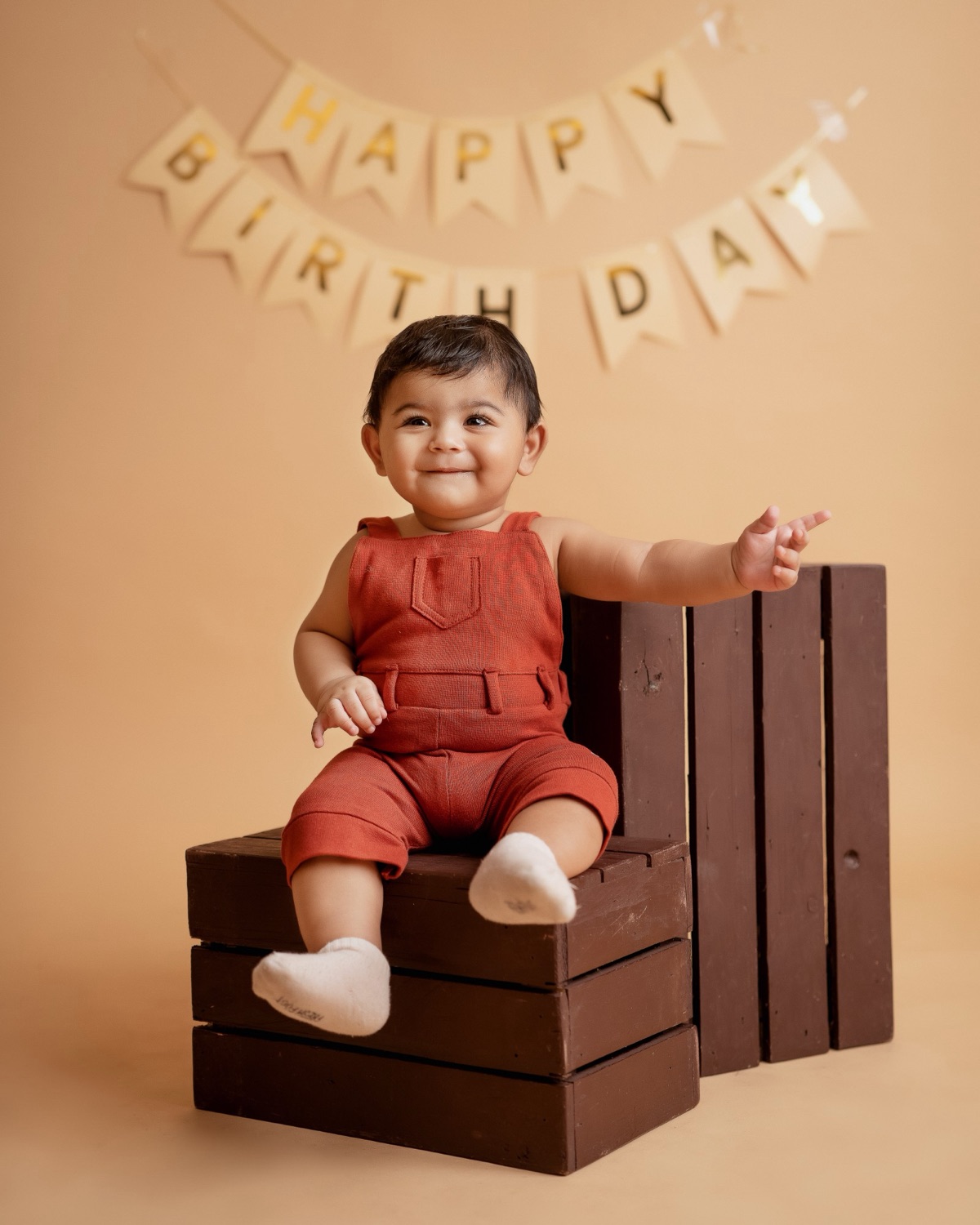 Baby in a rust romper on a wooden crate under a Happy Birthday banner