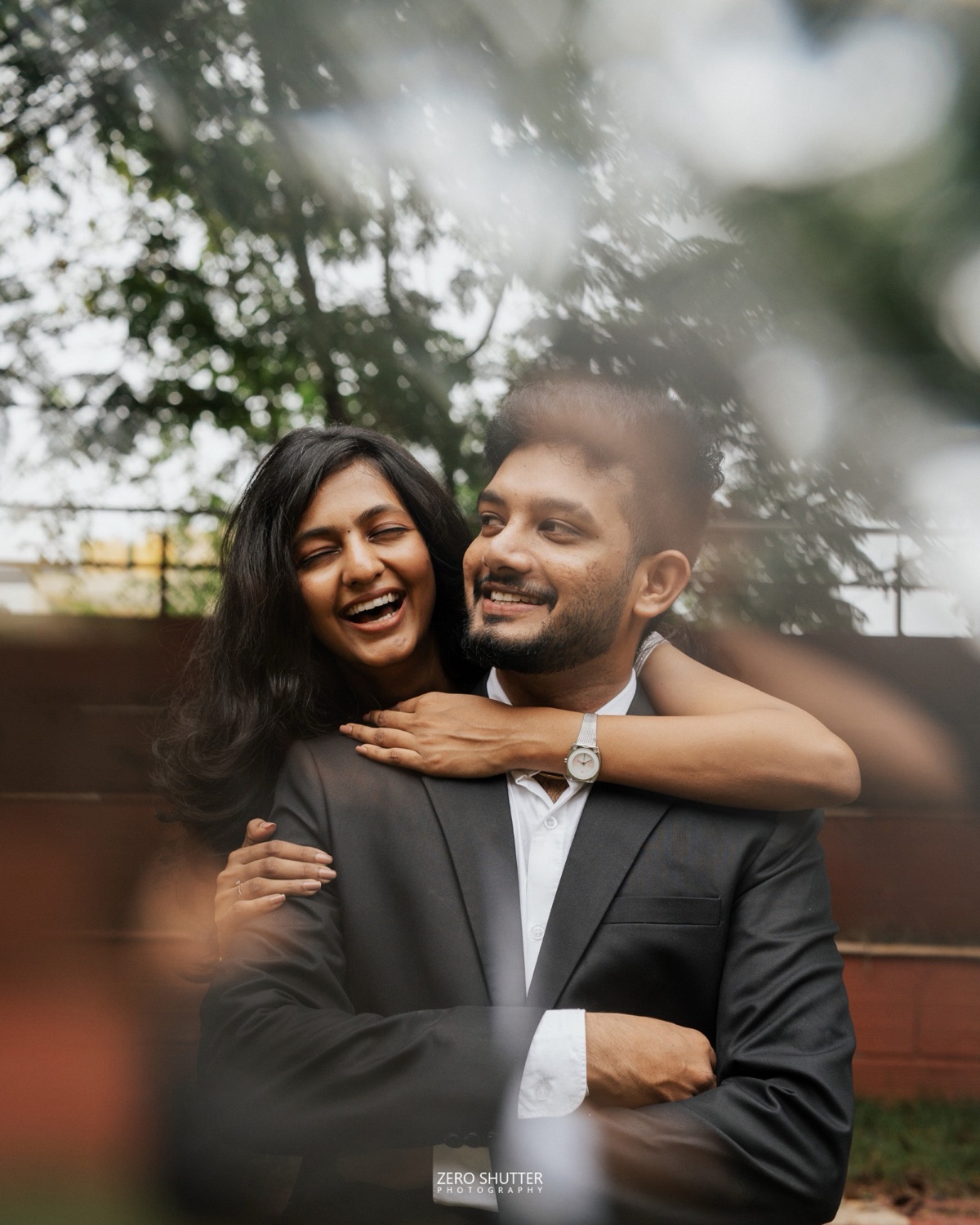 Wife laughing with her arms around her husband's shoulders, framed through soft out-of-focus foliage